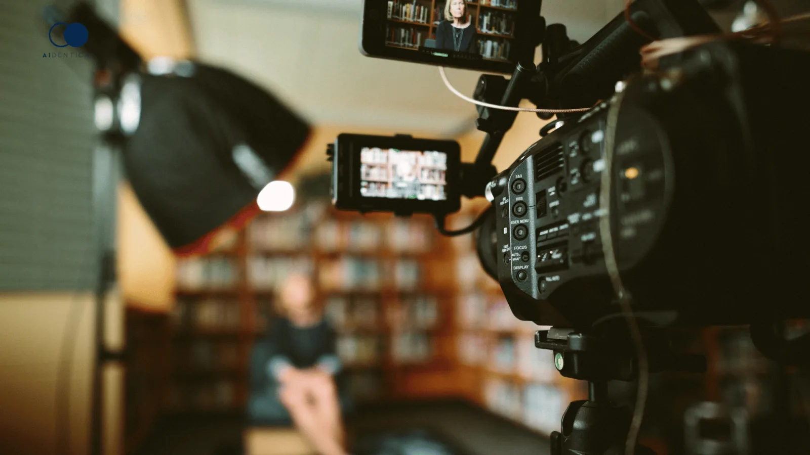 Filmkamera auf einem Stativ, dahinter unscharf eine sprechende Person im Aufnahmeraum